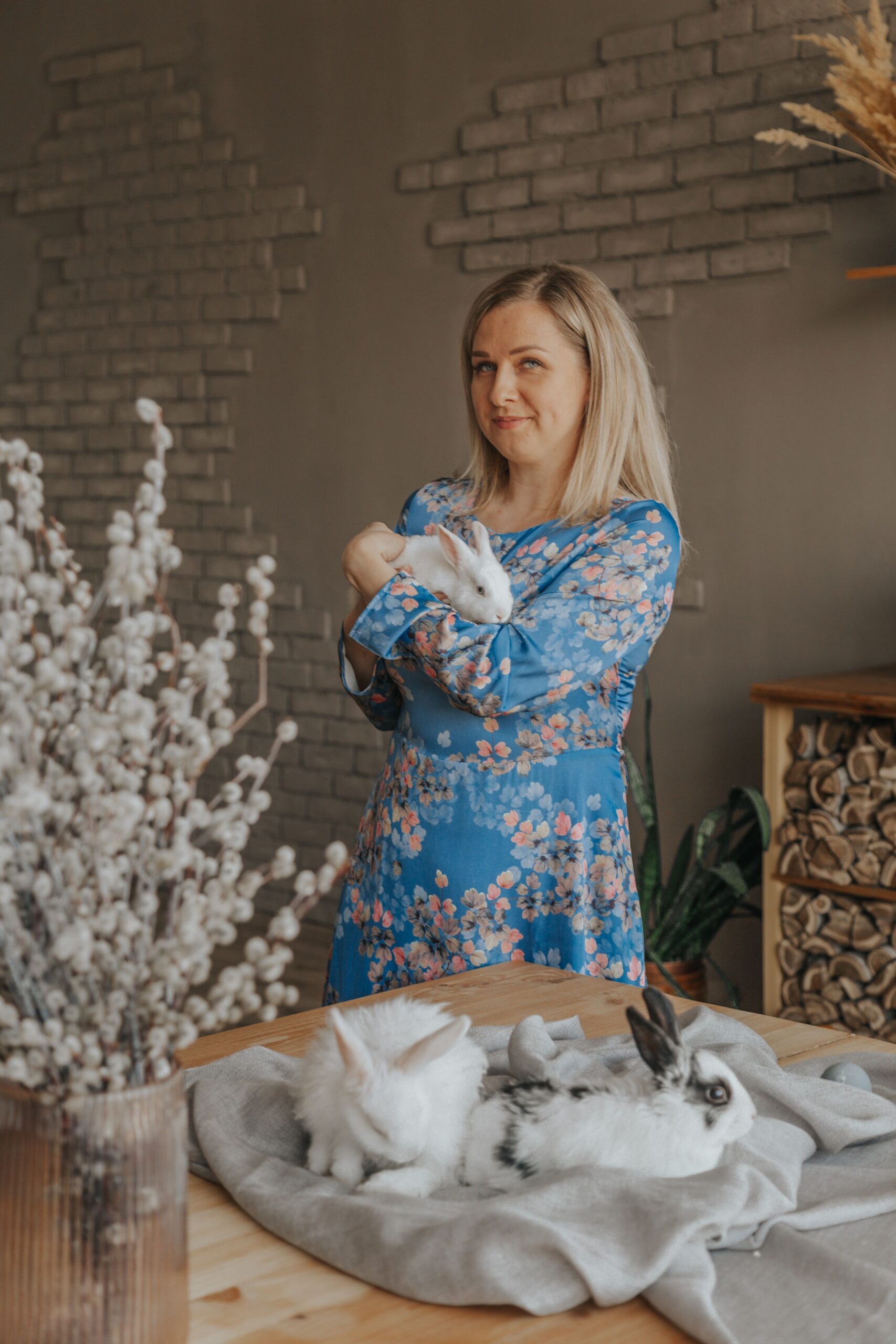 Woman in a blue floral dress holding a white rabbit, with two more rabbits resting on a table